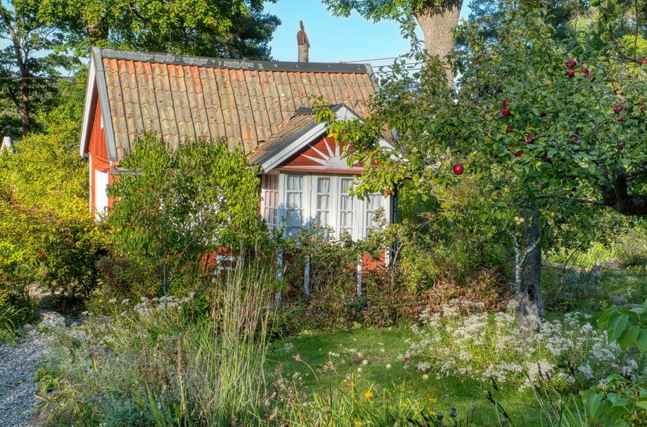 A quaint small cottage garden featuring a red wooden house with a tiled roof and a prominent white bay window. In the foreground, lush green grass and a variety of flowering plants and shrubs are visi