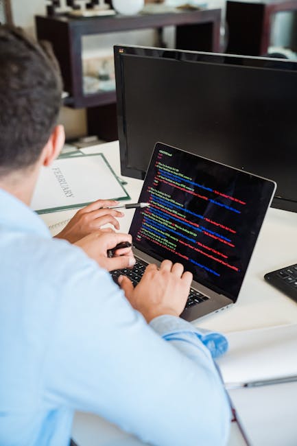 A person working at a desk outdoors, examining a laptop displaying lines of colorful code. The workspace features a computer monitor, a notebook, and a smartphone. In the background, a garden with nea